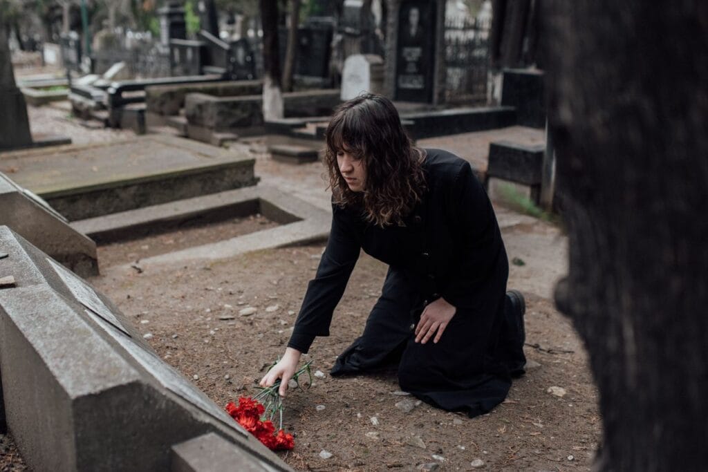 A woman in black kneels in a cemetery placing flowers on a grave.